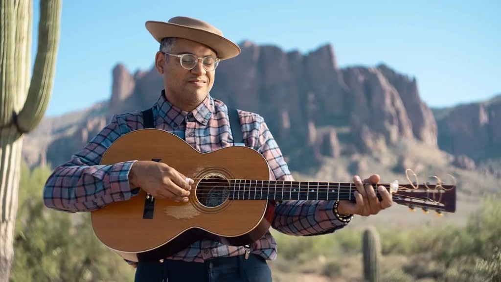 Dom Flemons playing acoustic guitar in the desert with mesa and cactus in the background